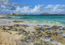 Fuerteventura, trá charraigeach ar fharraige thuircíse, le hóstán bán agus oileáin bholcánacha sa chúlra.