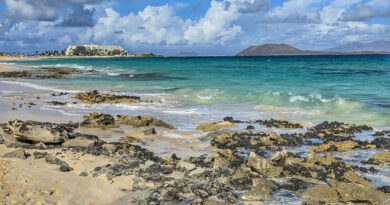 Fuerteventura, skalista plaża nad turkusowym morzem, z białym hotelem i wulkanicznymi wyspami w tle.