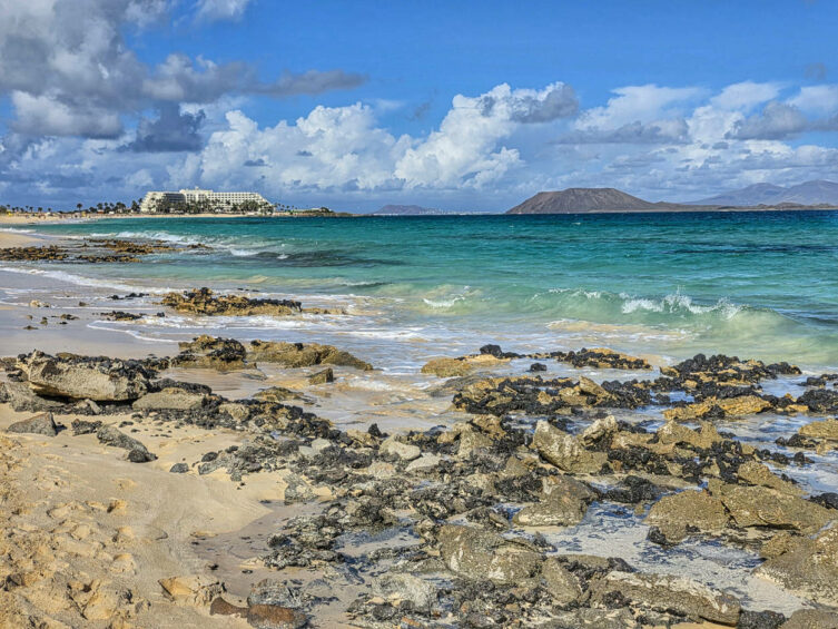 Fuerteventura, skalista plaża nad turkusowym morzem, z białym hotelem i wulkanicznymi wyspami w tle.