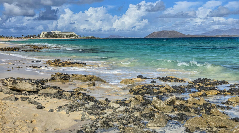Fuerteventura, skalista plaża nad turkusowym morzem, z białym hotelem i wulkanicznymi wyspami w tle.